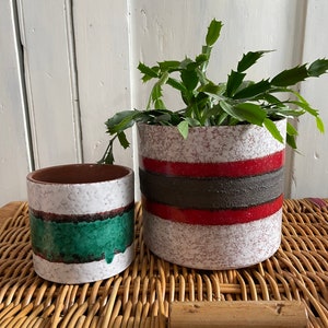 May include: Two ceramic plant pots with a white speckled glaze and colourful stripes. The larger pot has red, black, and grey stripes. The smaller pot has green and white stripes. Both pots are sitting on a woven wicker surface.