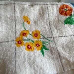 Op de afbeelding: Close-up van een witte, getextureerde stof met een rasterpatroon. Op de stof zijn gele bloemen met rode centra en groene bladeren geborduurd. Een oranje bloem met groene bladeren is ook zichtbaar.