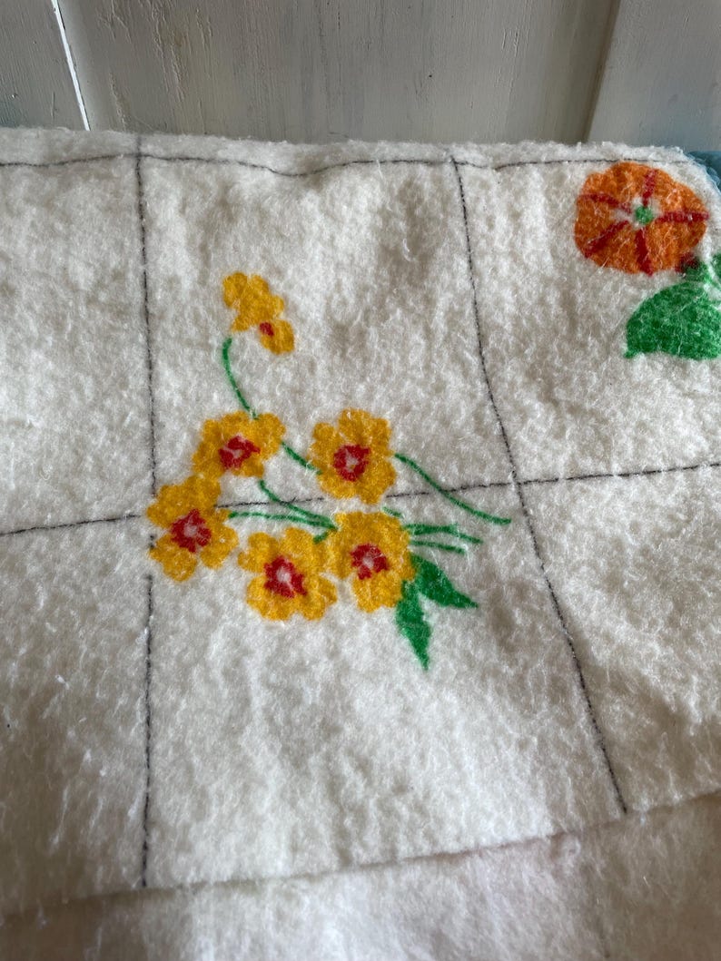 Op de afbeelding: Close-up van een witte, getextureerde stof met een rasterpatroon. Op de stof zijn gele bloemen met rode centra en groene bladeren geborduurd. Een oranje bloem met groene bladeren is ook zichtbaar.
