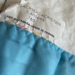 Op de afbeelding: Close-up van een label met de tekst "6% ACRYLIC-ACRYLIQUE CA00692 165 FAIT AU CANADA MADE IN CANADA" over een lichtblauwe stof met een gewatteerd patroon. De achtergrond is een witte, getextureerde ondergrond.