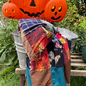May include: A patchwork quilt featuring a mix of fabrics and colours, including blue, red, brown, and green. The quilt is hanging on a metal bucket and is partially obscured by three orange jack-o'-lantern pumpkins.