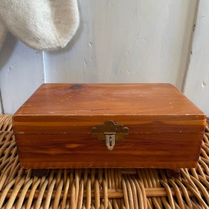May include: A wooden box with a brass latch. The box is made of cedar wood and has a natural wood finish. The box is sitting on a woven wicker surface.