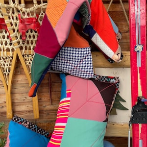 Peut inclure: Plusieurs coussins colorés en patchwork et une courtepointe pliée. Les coussins présentent des motifs géométriques roses, turquoise et bleus, avec un motif pied-de-poule. Une paire de skis rouges vintage et une raquette en bois sont en arrière-plan.