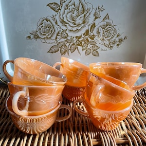 Op de afbeelding: Een stapel van zes vintage perzikkleurige glazen theekopjes met een bloemenmotief. De kopjes zijn gestapeld op een geweven rieten oppervlak.