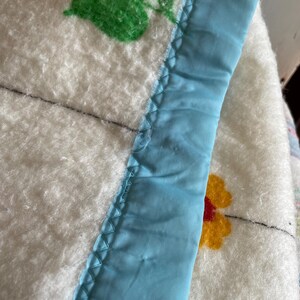 Op de afbeelding: Close-up van een witte deken met bloemmotieven in oranje, geel en groen. De deken heeft een lichtblauwe satijnen rand met een zigzagsteek. De deken lijkt vintage te zijn.