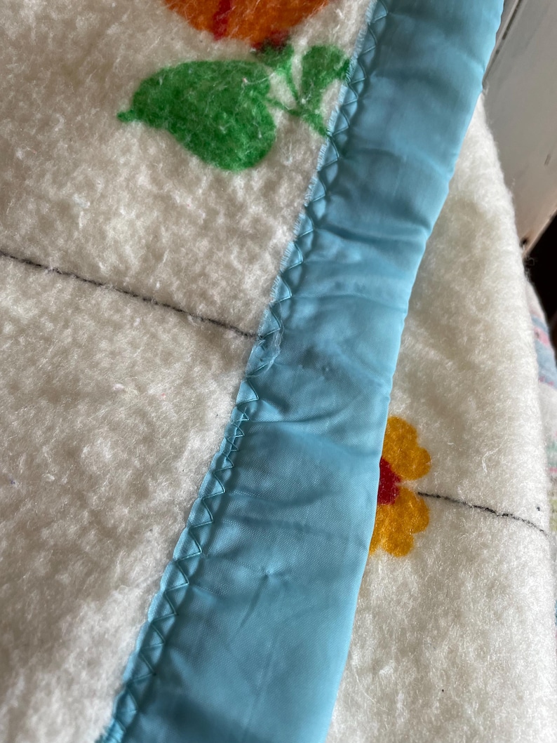 Op de afbeelding: Close-up van een witte deken met bloemmotieven in oranje, geel en groen. De deken heeft een lichtblauwe satijnen rand met een zigzagsteek. De deken lijkt vintage te zijn.