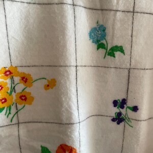 Op de afbeelding: Witte stof met een rasterpatroon en kleurrijke bloemmotieven. De bloemen zijn in tinten oranje, geel, blauw en paars, met groene bladeren. De rasterlijnen zijn zwart.