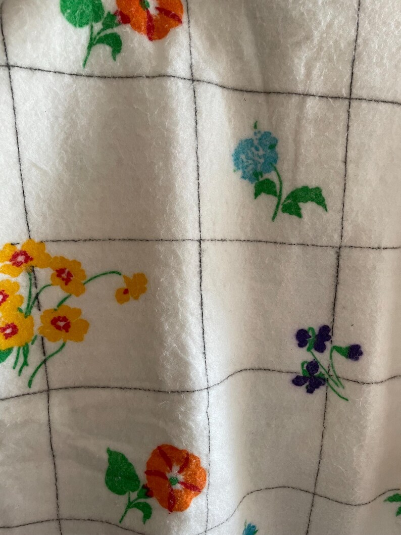 Op de afbeelding: Witte stof met een rasterpatroon en kleurrijke bloemmotieven. De bloemen zijn in tinten oranje, geel, blauw en paars, met groene bladeren. De rasterlijnen zijn zwart.