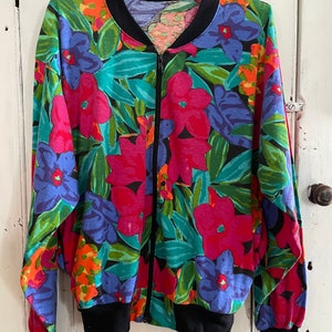 Op de afbeelding: Een zwart ritsjack met een kleurrijke bloemenprint. De print heeft grote roze, blauwe en oranje bloemen op een donkergroene achtergrond.