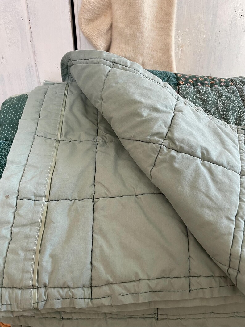 K&ouml;nnte beinhalten: Eine gesteppte Tagesdecke in Salbeigr&uuml;n mit einem wei&szlig;en Rei&szlig;verschlussdetail. Die Decke ist in der H&auml;lfte gefaltet und der Rei&szlig;verschluss ist an der Seite sichtbar.