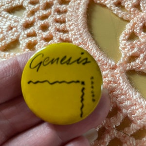 May include: Yellow round pinback button with black text that reads "Genesis" and a black squiggly line design.