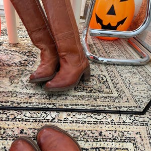 May include: A pair of knee-high brown leather boots with a stacked heel and subtle decorative stitching along the shaft. The boots feature a rounded toe and a smooth finish, reflecting in a mirror surface that also shows a plastic orange jack-o'-lantern.
