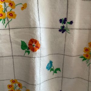 Op de afbeelding: Witte stof met een rasterpatroon en kleurrijke bloemborduurwerken. De bloemen zijn geel, oranje, blauw en paars, met groene bladeren. De stof heeft een zachte, getextureerde uitstraling.