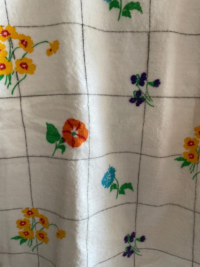 Op de afbeelding: Witte stof met een rasterpatroon en kleurrijke bloemborduurwerken. De bloemen zijn geel, oranje, blauw en paars, met groene bladeren. De stof heeft een zachte, getextureerde uitstraling.