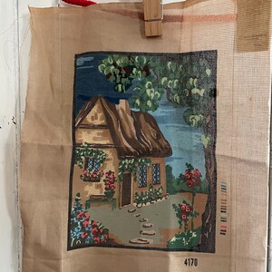 Peut inclure: Une tapisserie au point de croix vintage représentant une scène de cottage avec un toit de chaume, des fleurs et de la verdure. La tapisserie en toile beige est maintenue par une pince à linge en bois et une corde rouge. Le numéro "4170" est visible.