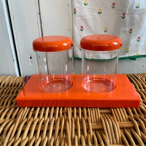 Peut inclure: Deux bocaux en verre transparent avec des couvercles orange sur un plateau orange vif. Les bocaux sont posés sur une surface en osier tressé.