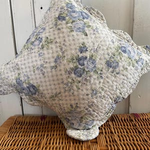 Peut inclure: Un coussin matelassé avec un motif floral de roses bleues et de feuilles vertes sur un fond à carreaux blancs et gris clair. Le coussin a un bord festonné et est posé sur un panier en osier.