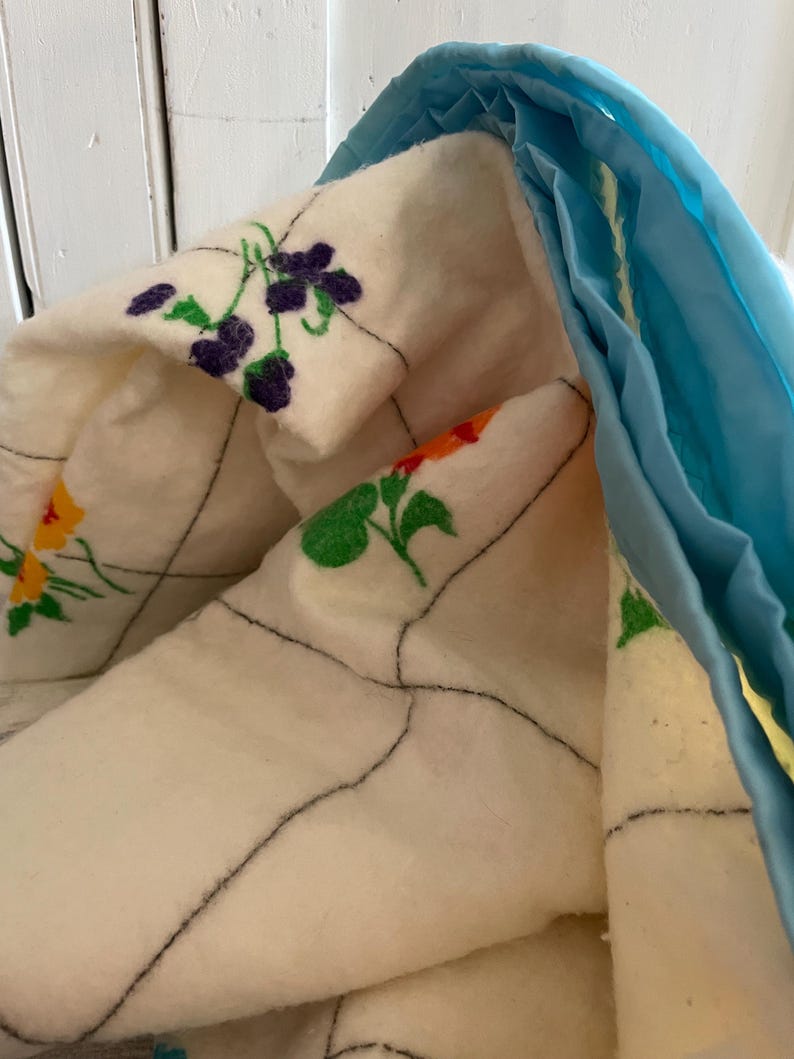 Op de afbeelding: Close-up van een gewatteerde deken met bloemenborduurwerk. De deken heeft een cr&egrave;mekleurige basis met zwarte stiksels en geborduurde bloemen in paars, oranje en geel. Een lichtblauwe stof omzoomt de deken.