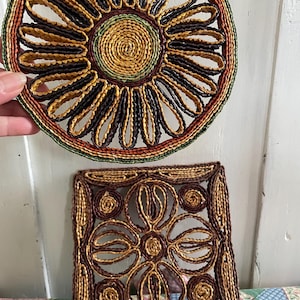 May include: Two woven trivets. The round trivet has a sunflower design with a yellow centre, surrounded by brown and black loops. The square trivet features a floral pattern in brown and tan. Both are made of natural fibres.