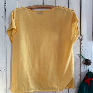 Op de afbeelding: Een geel shirt met korte mouwen, een ronde hals en knopen op de schouders.
