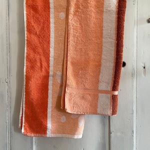 Op de afbeelding: Twee oranje en witte gestreepte handdoeken hangen aan een houten hanger. De handdoeken hebben een witte rand en een bruine streep aan de rand.