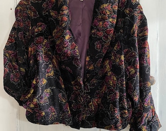 May include: A black velvet jacket with a floral pattern in shades of purple, gold, and green. The jacket has a shawl collar and long sleeves. The label "Le Chateau" is visible on the inside of the jacket.