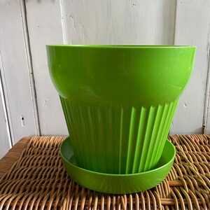 May include: A bright green plastic plant pot with a matching saucer. The pot has a fluted design and is sitting on a woven wicker surface.