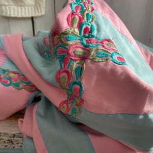 Peut inclure: Un textile plié avec des panneaux alternés bleu clair et rose. Les bords sont ornés de motifs de plumes de paon brodés en rose, vert et turquoise. Le tissu semble doux et fluide.