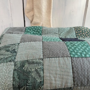 Peut inclure: Une courtepointe patchwork avec une variété de motifs verts et gris. La courtepointe est faite de carrés de tissus différents, y compris du vichy, des imprimés floraux et géométriques. La courtepointe est posée sur un panier en osier.