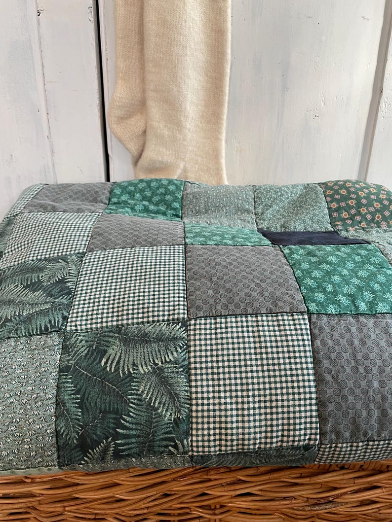 K&ouml;nnte beinhalten: Eine Patchwork-Decke mit verschiedenen gr&uuml;nen und grauen Mustern. Die Decke besteht aus Quadraten aus verschiedenen Stoffen, darunter Gingham, Blumen- und geometrische Drucke. Die Decke liegt auf einem Weidenkorb.
