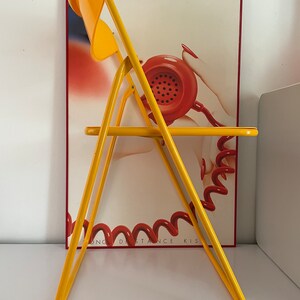 May include: A yellow folding chair with a curved back and a seat. The chair is in front of a poster with a red phone and the text "Long Distance Kiss".