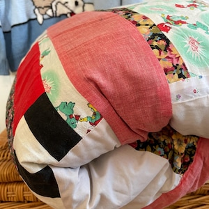 Peut inclure: Un coussin rond en patchwork avec divers tissus rouges, noirs, blancs et à motifs floraux. Le coussin repose sur un panier en osier. Un pull bleu clair avec un personnage de dessin animé est en arrière-plan.