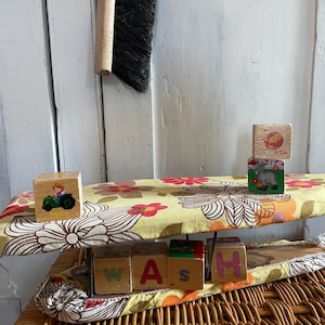 Peut inclure: Un planche à repasser jaune et marron à fleurs avec un cadre en métal. Le plateau est recouvert d'un tissu à motif floral. Il y a des blocs de bois qui épellent le mot "WASH" sur le plateau. Les blocs sont décorés avec des images d'un tracteur, d'une lettre A, d'une lettre S et d'une lettre H.