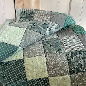 K&ouml;nnte beinhalten: Eine Patchwork-Decke aus verschiedenen gr&uuml;nen und grauen Stoffen. Die Decke besteht aus Quadraten mit unterschiedlichen Mustern, darunter Farne, Gingham und Punkte.
