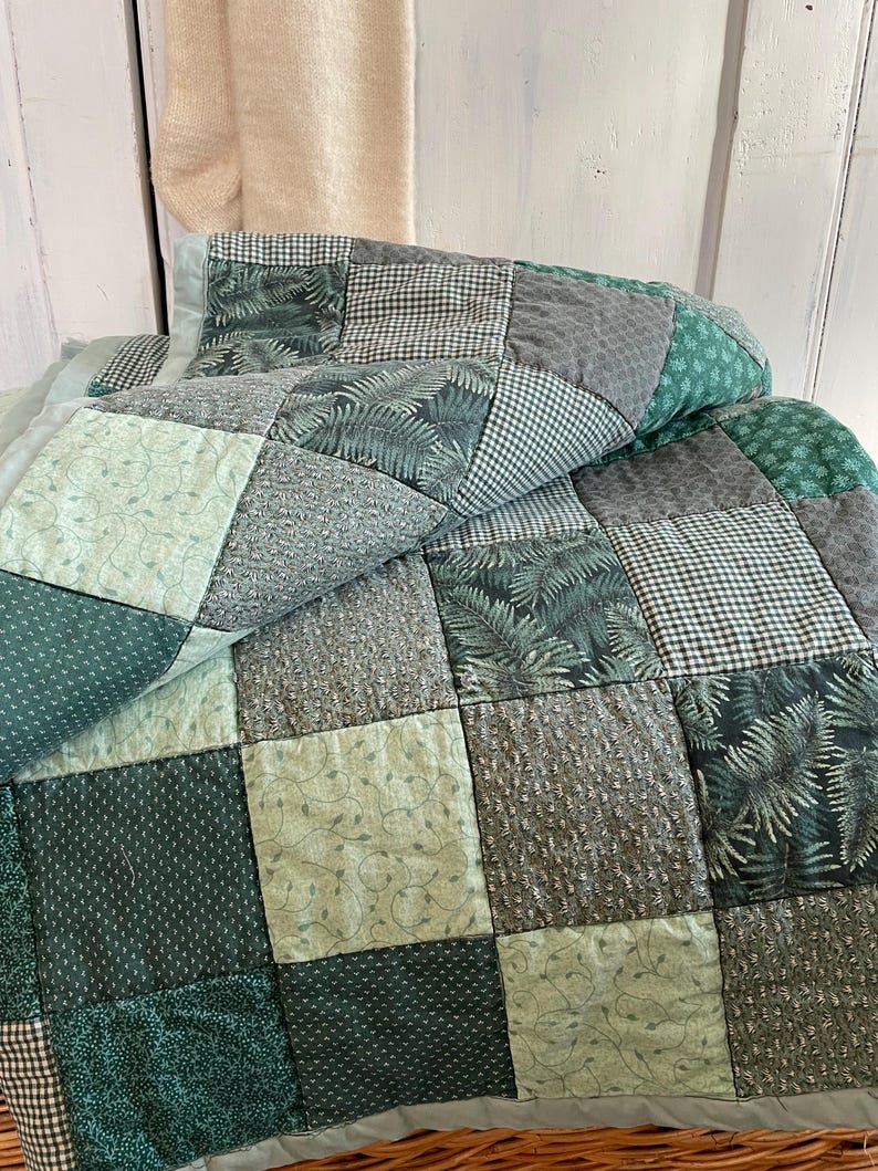 K&ouml;nnte beinhalten: Eine Patchwork-Decke aus verschiedenen gr&uuml;nen und grauen Stoffen. Die Decke besteht aus Quadraten mit unterschiedlichen Mustern, darunter Farne, Gingham und Punkte.