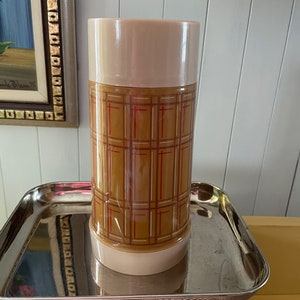 May include: A brown and white plaid flask with a cream-coloured lid. The flask is sitting on a silver scale that reads "for Trade".