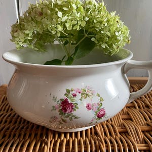 May include: White ceramic chamber pot with a floral design featuring pink and white roses. The pot has a handle on the side and is sitting on a woven wicker surface.