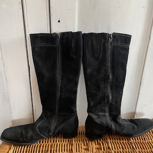 May include: A pair of black suede knee-high boots with side zips. The boots have a rounded toe and a low heel. The boots are displayed on a woven wicker surface against a white wooden background.