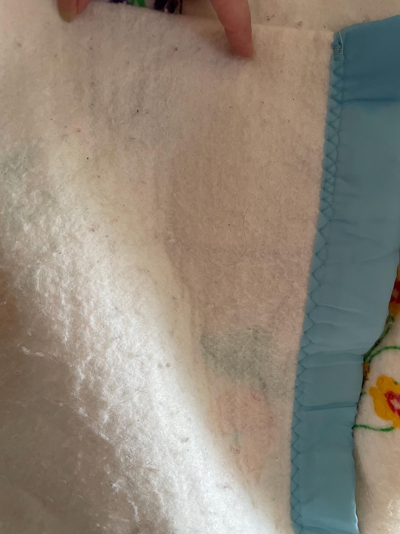 Op de afbeelding: Close-up van een wit gewatteerd deken met een lichtblauwe satijnen rand. De deken heeft een zacht, getextureerd oppervlak en een decoratief bloemenpatroon zichtbaar in de hoek.