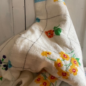Op de afbeelding: Een vintage witte deken met een bloemenpatroon in geel, oranje en paars. De deken heeft een blauwe rand en een rasterpatroon gestikt met zwarte draad. De deken is opgevouwen en gedrapeerd.