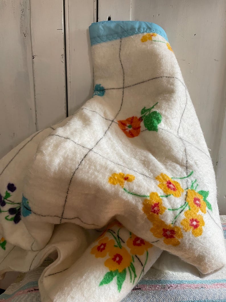 Op de afbeelding: Een vintage witte deken met een bloemenpatroon in geel, oranje en paars. De deken heeft een blauwe rand en een rasterpatroon gestikt met zwarte draad. De deken is opgevouwen en gedrapeerd.