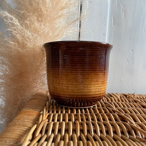 May include: A brown ceramic planter with a light brown glaze. The planter has a rounded shape and a slightly flared lip. It is sitting on a woven wicker surface.
