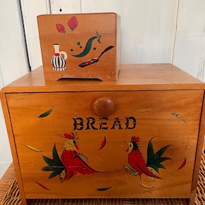 Peut inclure: Une boîte à pain en bois avec un motif de coq peint sur le devant. La boîte a un couvercle à charnières et un petit bouton sur le dessus. Les mots "BREAD" sont peints sur le devant de la boîte. Il y a une plus petite boîte en bois avec un motif peint de fraises, un pichet et une poêle à frire sur le dessus de la boîte à pain.