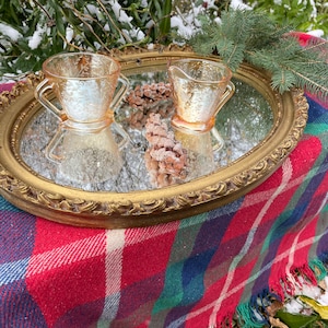 Peut inclure: Un plateau miroir ovale doré avec un cadre orné surélevé repose sur une couverture à carreaux rouge et vert. Deux crémiers et sucriers en verre vintage à motif floral sont sur le plateau.