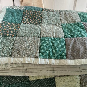K&ouml;nnte beinhalten: Eine gr&uuml;ne und graue Patchwork-Decke mit verschiedenen Blumenmustern. Die Decke ist in der H&auml;lfte gefaltet und hat einen hellgrauen Rand.