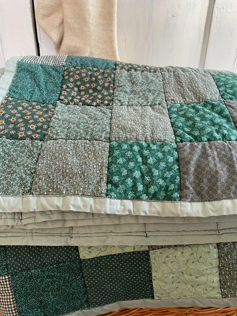 K&ouml;nnte beinhalten: Eine gr&uuml;ne und graue Patchwork-Decke mit verschiedenen Blumenmustern. Die Decke ist in der H&auml;lfte gefaltet und hat einen hellgrauen Rand.