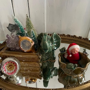 Peut inclure: Un miroir ovale doré présente une collection de figurines vintage. Un éléphant en céramique verte se tient à côté d'un contenant doré contenant des mini sapins de Noël, des raisins et une horloge. Une figurine du Père Noël est assise dans un bol en céramique verte.