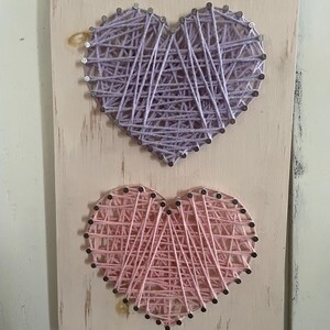 Peut inclure: Décoration murale en bois avec deux cœurs en fil de fer. Le cœur supérieur est lavande et le cœur inférieur est rose. Les deux cœurs sont faits de clous et de fil sur une planche de bois vieillie de couleur claire.