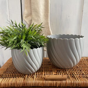 Peut inclure: Deux pots de fleurs en céramique gris avec un design ondulé. Le plus petit pot est rempli de plantes vertes et les deux pots sont posés sur une surface en osier tressé.