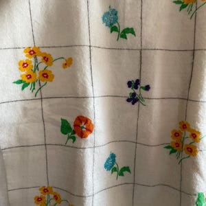 Op de afbeelding: Witte stof met een ruitpatroon, geborduurd met kleurrijke bloemen. De bloemen zijn in tinten van oranje, geel, blauw en paars, met groene bladeren. De stof lijkt zacht en getextureerd.